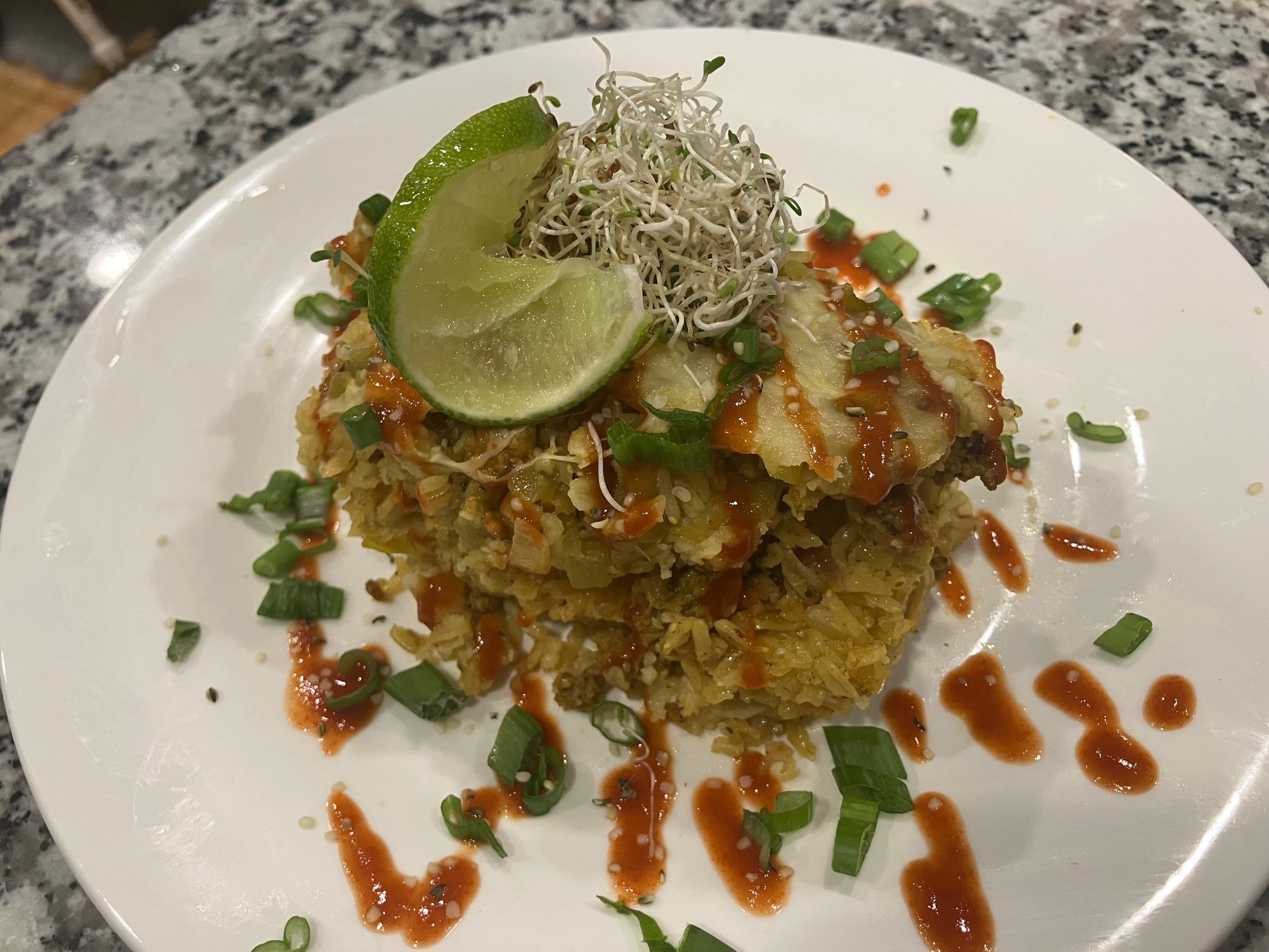 Seasoned rice topped with sprouts, a lime wedge, green onion and a red sauce drizzle on a white plate