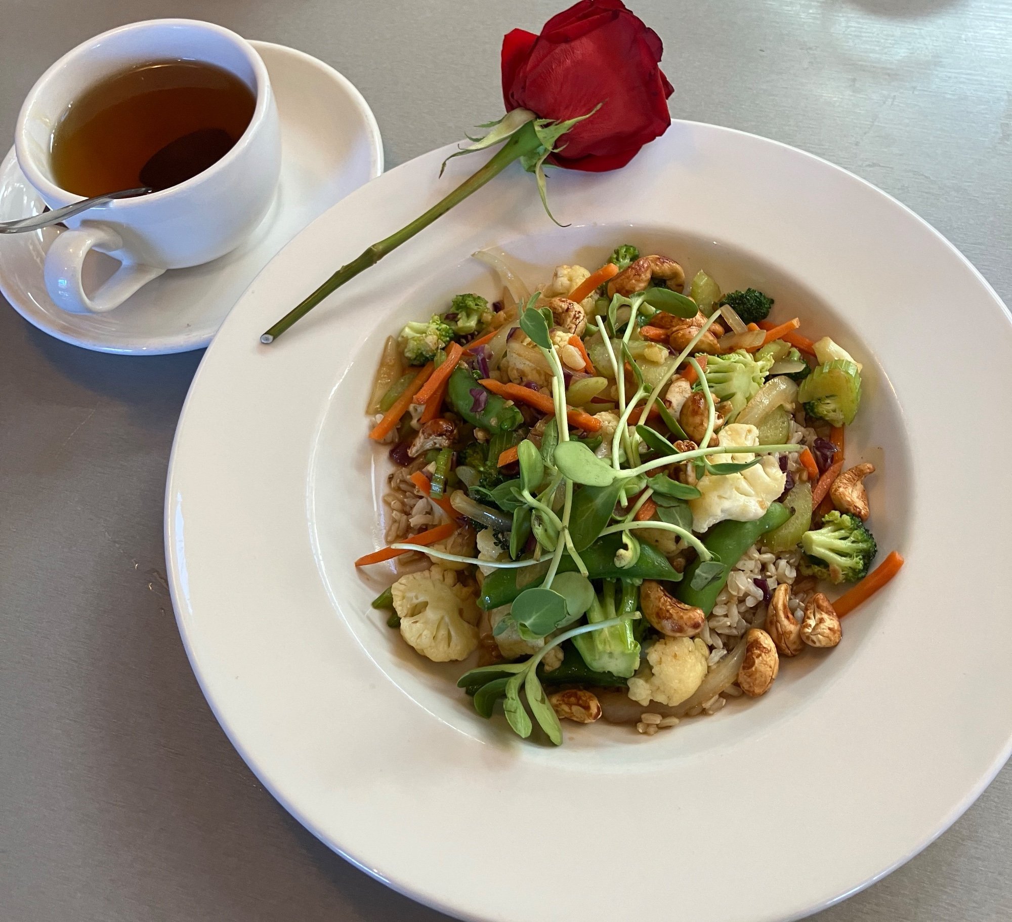 A bowl of stir-fried vegetables and cashews over brown rice, next to a cup of tea and a red rose