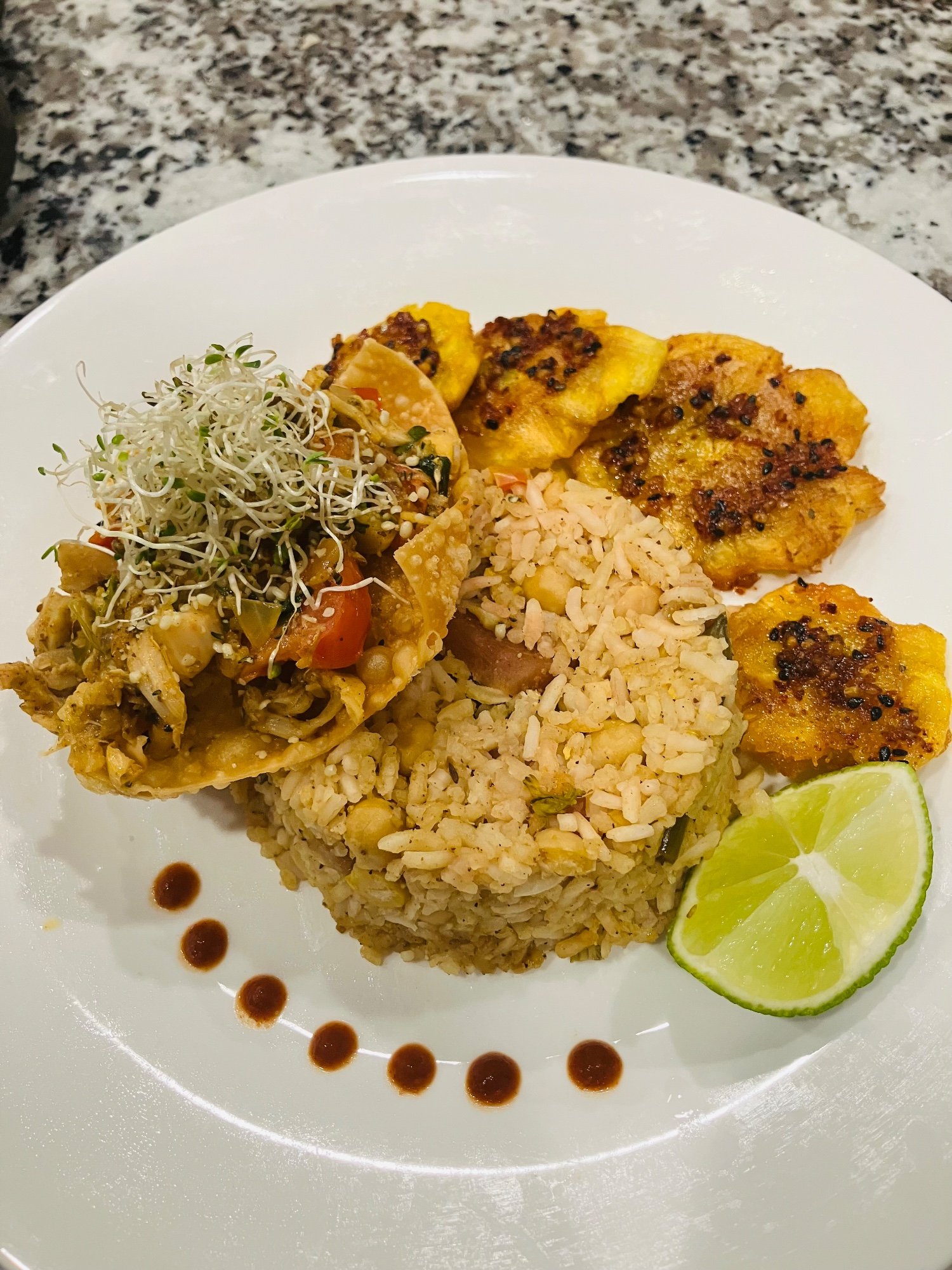 A plate of fried rice with chickpeas, crispy plantains, a sprout-topped filling and a lime wedge