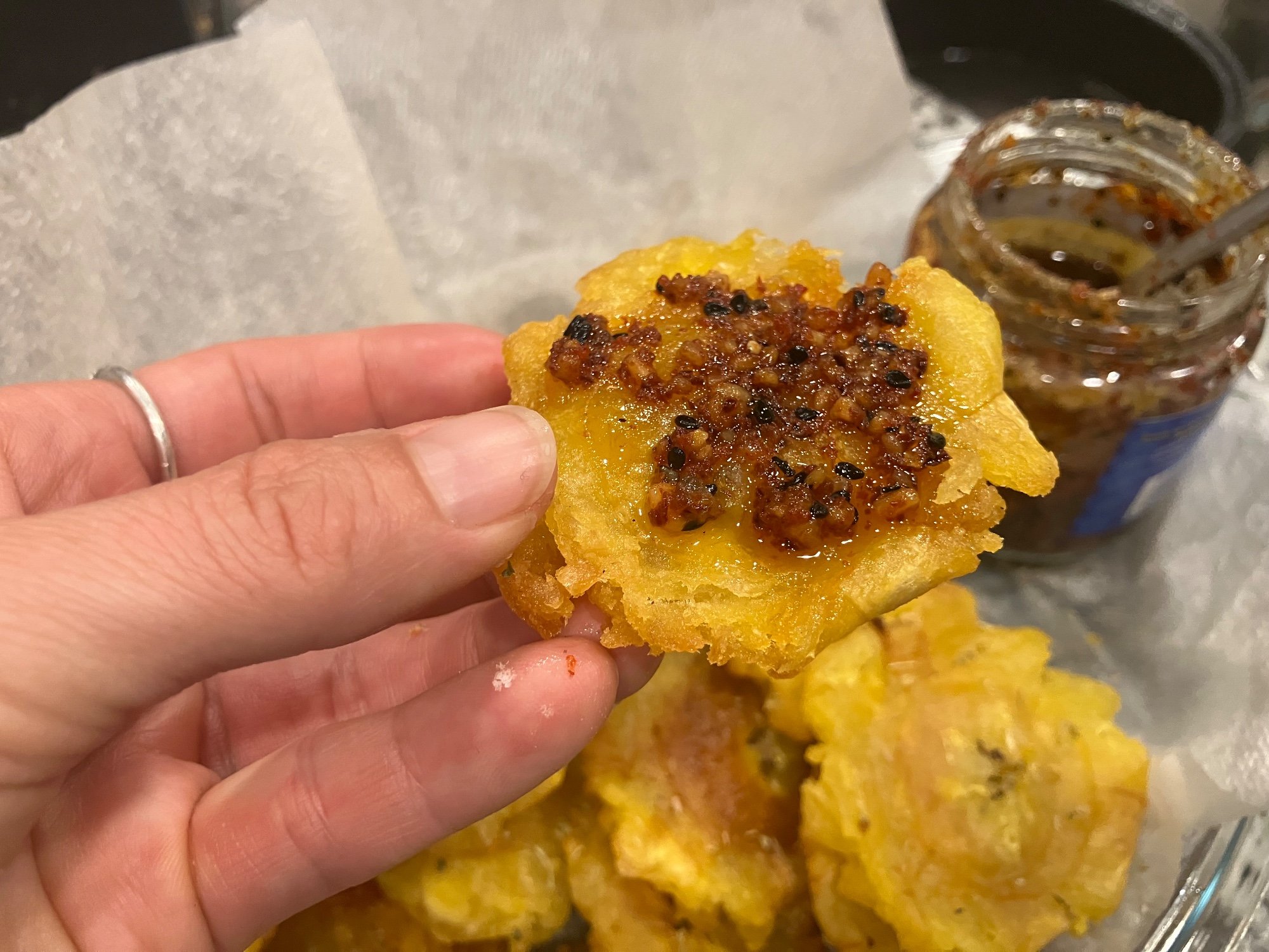 A hand holding a crispy fried plantain topped with a spicy chili crisp