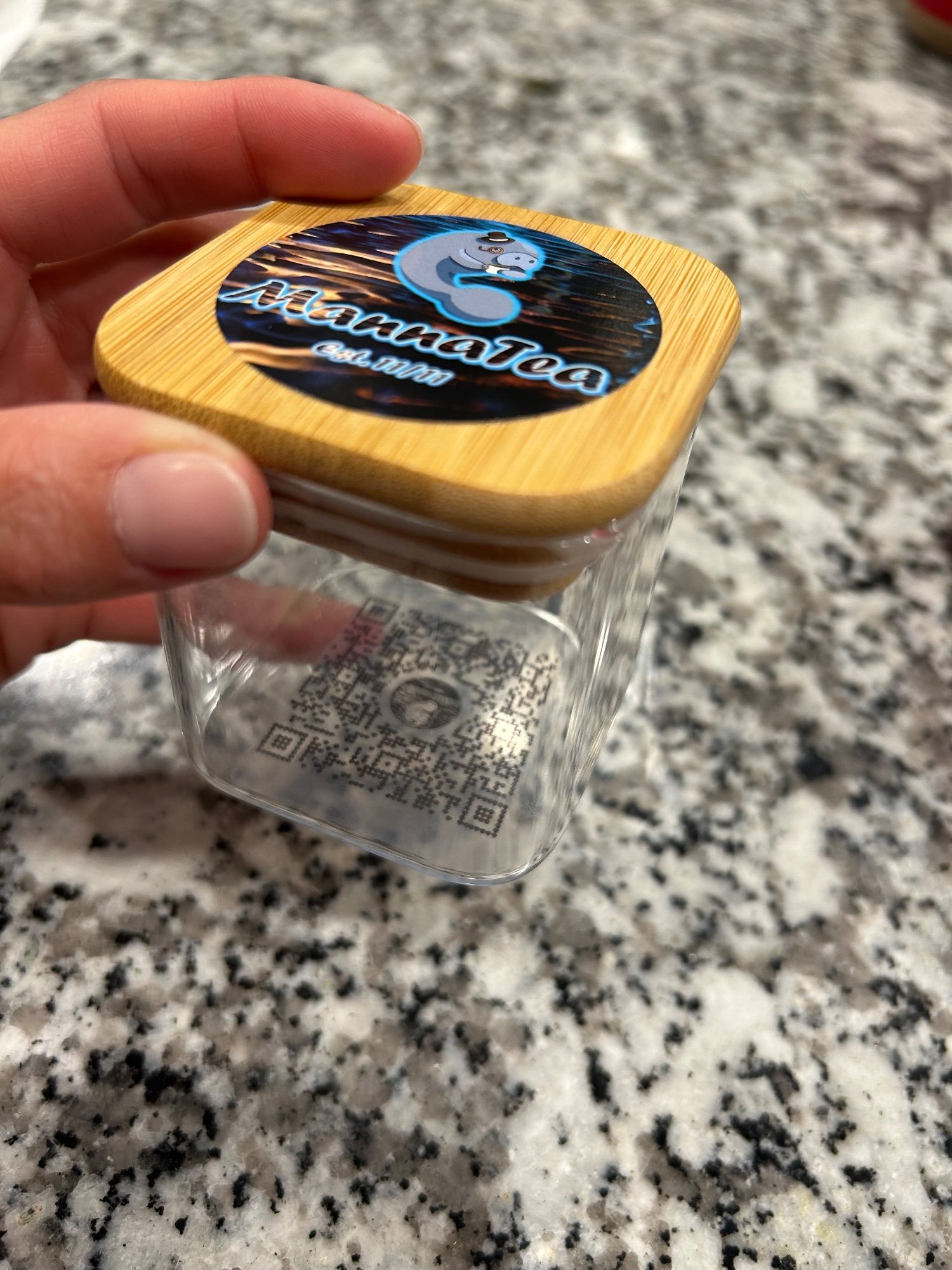 A hand holding a glass jar with a bamboo lid and a Manna Tea label