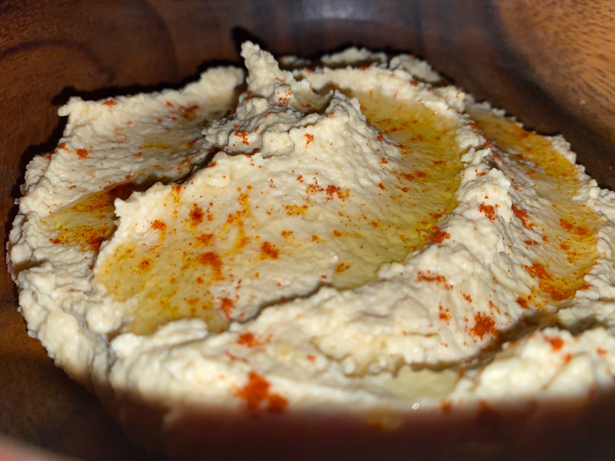 A wooden bowl of creamy hummus drizzled with olive oil and dusted with paprika