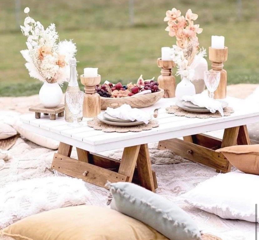 A low picnic table set with a wooden bowl of fresh fruit, candles, dried flowers and pillows on a blanket