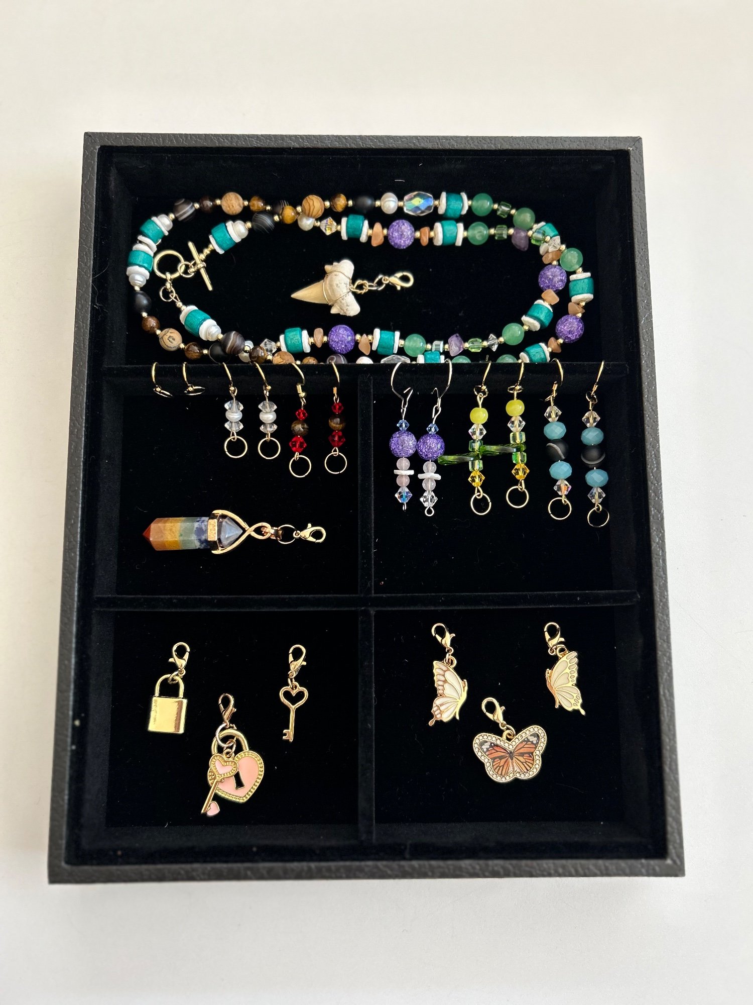 A black jewelry tray with beaded necklaces, earrings, and heart, key, lock and butterfly charms