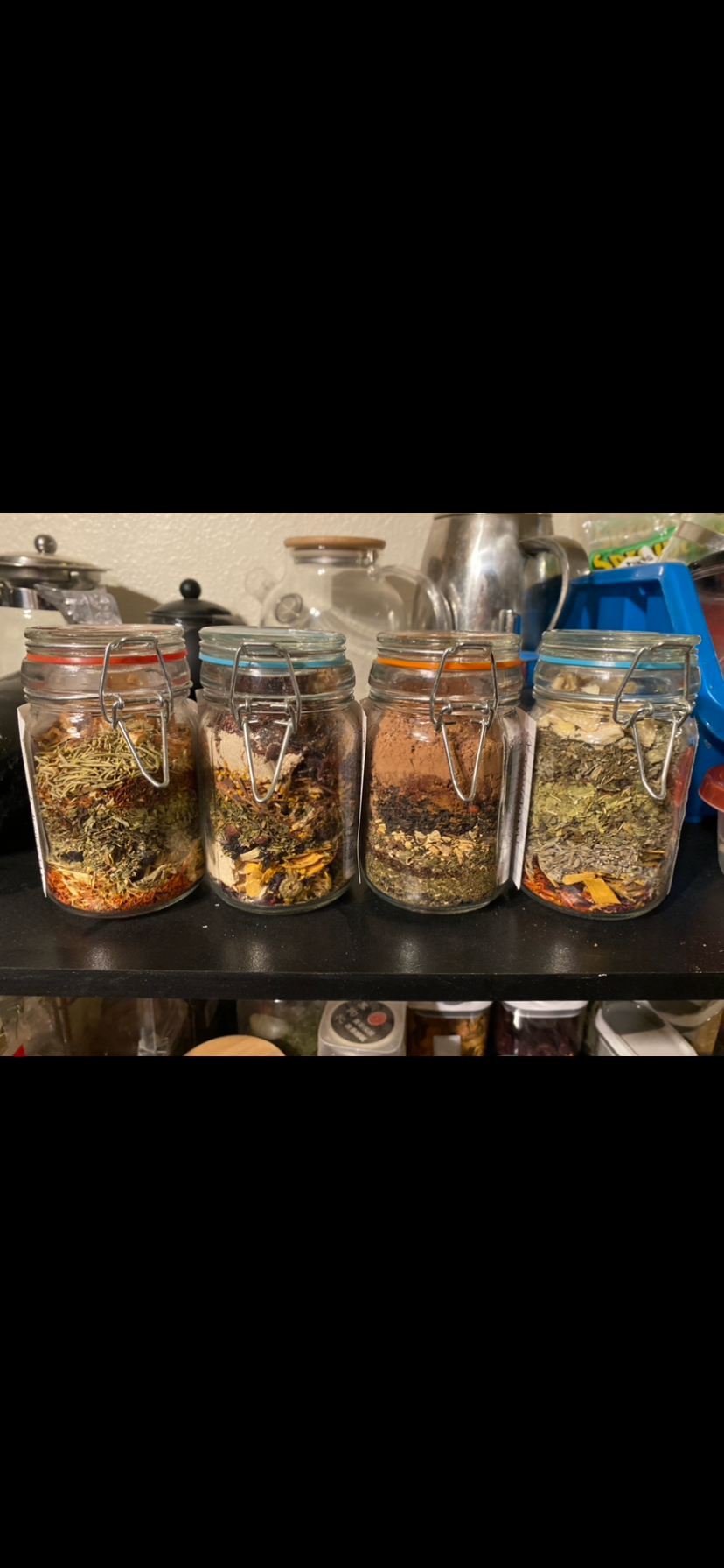 Four glass clip-top jars of loose herbal tea blends in a row on a shelf, each with a different colored band: red, blue, orange and teal