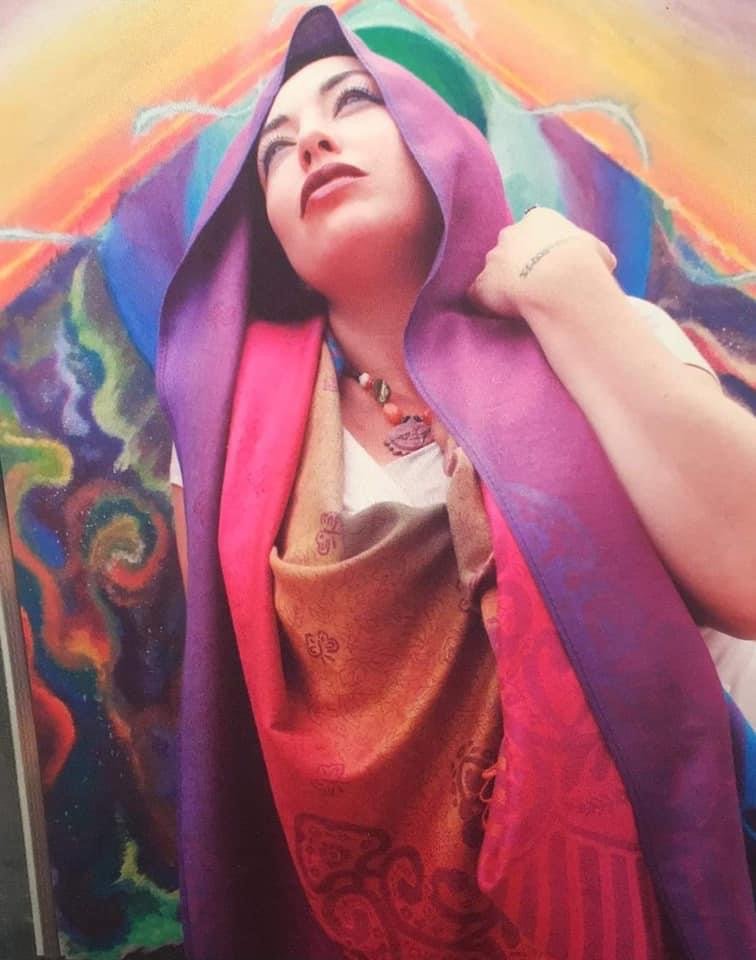 A woman draped in a hooded Karrma wrap in purple, pink and gold, standing in front of a colorful painted mural