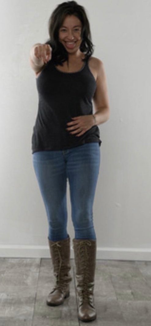 A smiling woman in a black tank top, jeans and brown lace-up boots, pointing toward the camera
